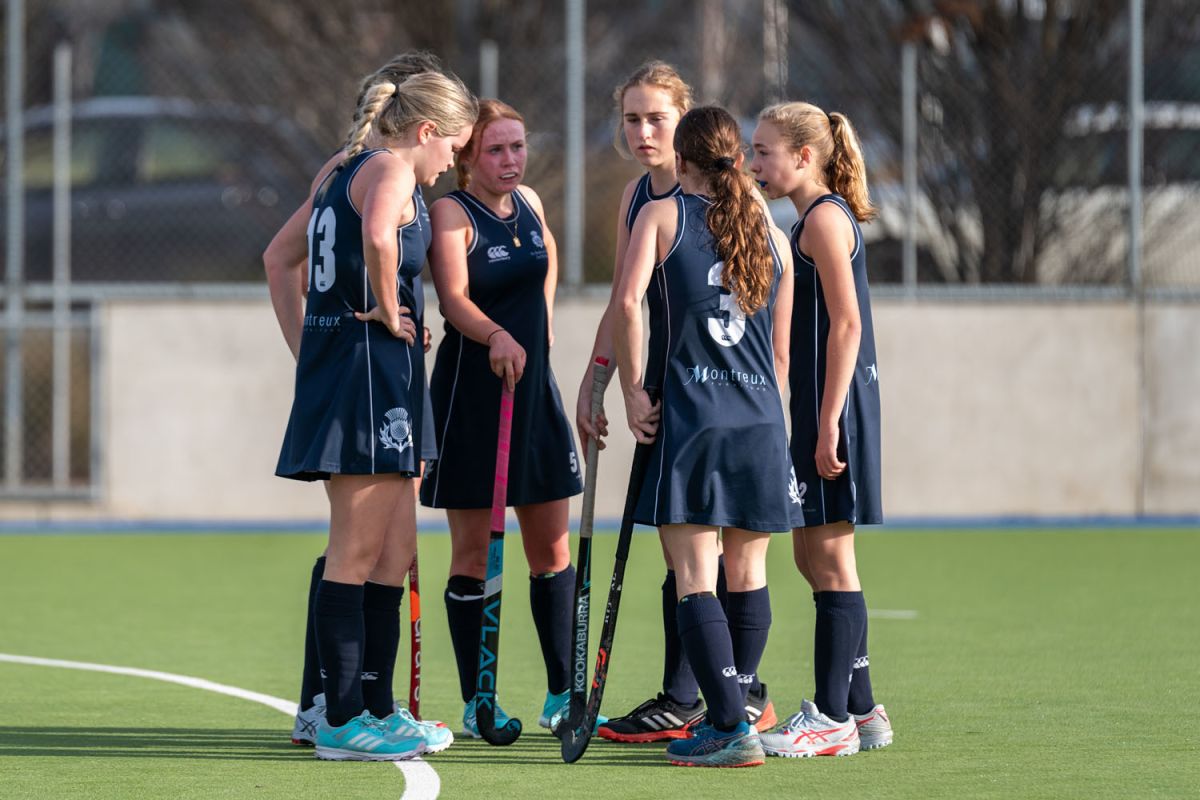 St Andrew's College female hockey players in sports uniform talking in a huddle on the hockey pitch.