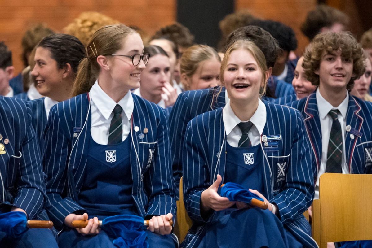 St Andrew's College students in uniform sat smiling and talking in chapel holding offering collection bags before service starts.